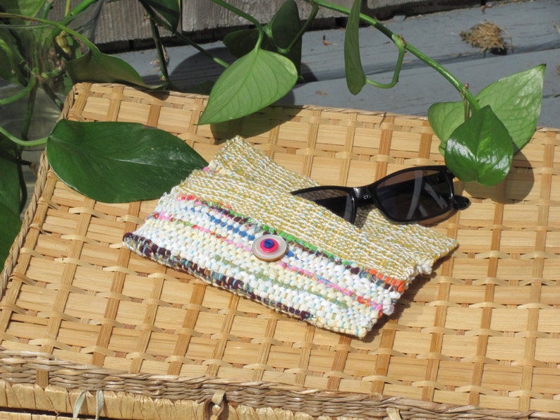 Boho Wallet Smart Phone Case, Small Hand Woven Clutch Purse, Cloth Fabric Soft Eyeglass Case Handmade Vegan Eco Friendly Zero Waste Upcycled Recycled Bag, Sustainable Slow Fashion Accessory with Sunny Yellow Accents Handcrafted by Amy C. Lund! Showing button closure. Styled open with sunglasses on a basket with green leaves.