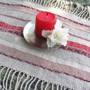 Handcrafted Small Positive Energy Red Altar Cloth Table Mat, Handmade Artisan Woven Spiritual Meditation, Reiki Root Chakra, Fire Element Feng Shui, Fire Red Fabric Table Centerpiece by Amy C. Lund in Burgundy Red, Blush Rose and Beige Gray with striped bands of alternating color and texture patterns. Styled on a rock surface with a red candle is sitting on a silver plate and white flower.