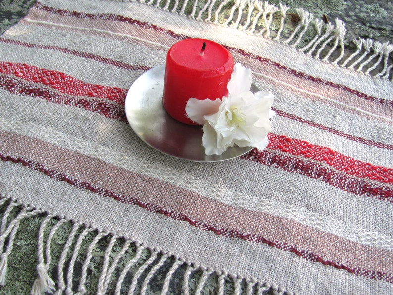 Handcrafted Small Positive Energy Red Altar Cloth Table Mat, Handmade Artisan Woven Spiritual Meditation, Reiki Root Chakra, Fire Element Feng Shui, Fire Red Fabric Table Centerpiece by Amy C. Lund in Burgundy Red, Blush Rose and Beige Gray with striped bands of alternating color and texture patterns. Styled on a rock surface with a red candle is sitting on a silver plate and white flower.