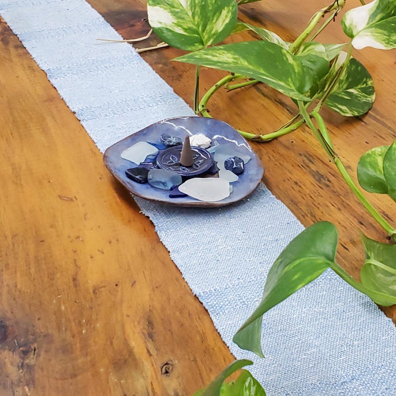 Artisan Handmade Light Blue Spring Summer Scarf, Minimalist Zen Serenity, Simplicity, Soothing Tranquil Tones Meditation Table Runner, Gender Neutral Unisex Men's or Women's Om Yoga, Spiritual Prayer Clothing, Hand Woven Cotton Scarf, Slow Fashion Accessory or Home Decor Accent Gift Idea. Styled on a wood table with a blue ceramic dish holding seaglass, shells, sodalite and an incense cone. Green plant leaves in background.