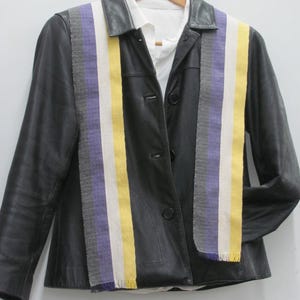 Puede incluir: Chaqueta de cuero negra con una camisa blanca debajo. La chaqueta presenta un diseño a rayas de colores en morado, amarillo y blanco. La chaqueta está colgada en una percha de madera.