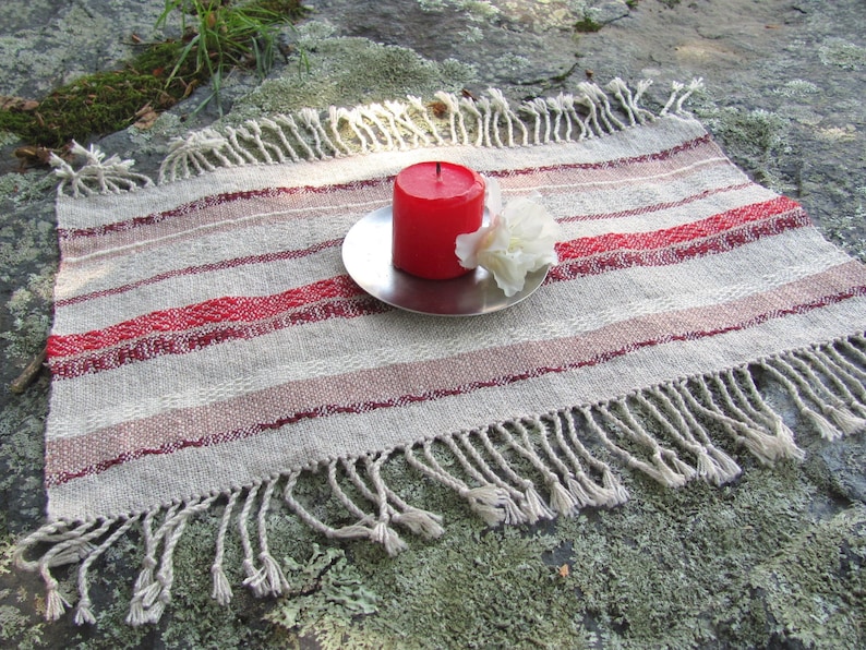 Handcrafted Small Positive Energy Red Altar Cloth Table Mat, Handmade Artisan Woven Spiritual Meditation, Reiki Root Chakra, Fire Element Feng Shui, Fire Red Fabric Table Centerpiece by Amy C. Lund in Burgundy Red, Blush Rose and Beige Gray with striped bands of alternating color and texture patterns. Styled on a rock surface with a red candle is sitting on a silver plate and white flower.