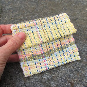 May include: A small woven pouch with a yellow, white, pink, blue, and gray pattern. The pouch is folded in half and has a flap.