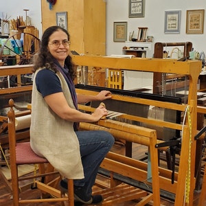 Amy C. Lund Handweaver and fiber artist, sitting at a loom in the weaving studio and gallery where she teaches and creates artisan handspun yarns from sheep's wool, alpaca, cotton, linen and other fibers; hand knit and hand woven accessories including scarves, handbags and clothing; home decor table linens, blankets, rugs, tapestries; mixed media fiber art collage assemblages; and repairs textiles. She has a M.S. degree in Textiles & Related Art from the University of Rhode Island in 1993.