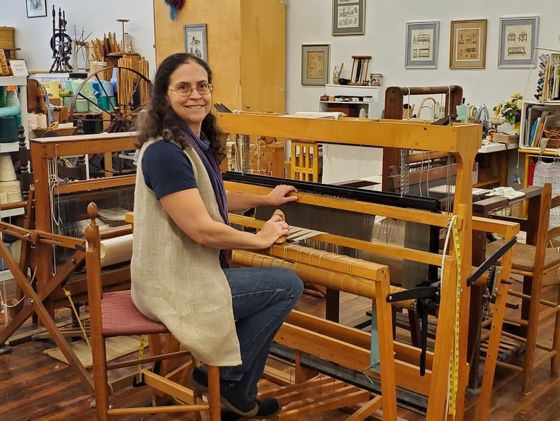 Amy C. Lund Handweaver and fiber artist, sitting at a loom in the weaving studio and gallery where she teaches and creates artisan handspun yarns from sheep's wool, alpaca, cotton, linen and other fibers; hand knit and hand woven accessories including scarves, handbags and clothing; home decor table linens, blankets, rugs, tapestries; mixed media fiber art collage assemblages; and repairs textiles. She has a M.S. degree in Textiles & Related Art from the University of Rhode Island in 1993.