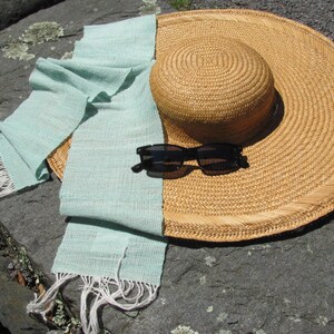 May include: A light blue woven scarf with white fringe lies on a gray stone surface with a large straw sun hat and black sunglasses.