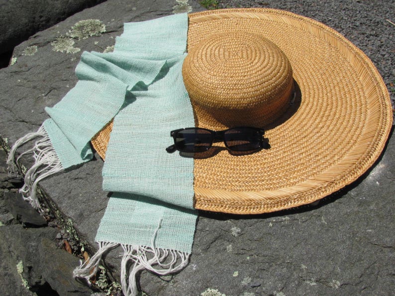 May include: A light blue woven scarf with white fringe lies on a gray stone surface with a large straw sun hat and black sunglasses.