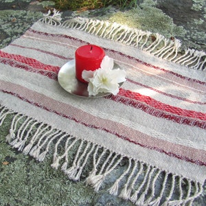 Handcrafted Small Positive Energy Red Altar Cloth Table Mat, Handmade Artisan Woven Spiritual Meditation, Reiki Root Chakra, Fire Element Feng Shui, Fire Red Fabric Table Centerpiece by Amy C. Lund in Burgundy Red, Blush Rose and Beige Gray with striped bands of alternating color and texture patterns. Styled on a rock surface with a red candle is sitting on a silver plate and white flower.