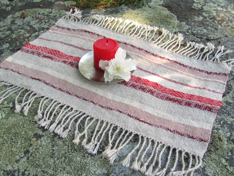 Handcrafted Small Positive Energy Red Altar Cloth Table Mat, Handmade Artisan Woven Spiritual Meditation, Reiki Root Chakra, Fire Element Feng Shui, Fire Red Fabric Table Centerpiece by Amy C. Lund in Burgundy Red, Blush Rose and Beige Gray with striped bands of alternating color and texture patterns. Styled on a rock surface with a red candle is sitting on a silver plate and white flower.