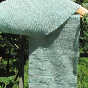 May include: A light blue woven fabric scarf hanging on a wooden hanger. The fabric has a textured, woven pattern.
