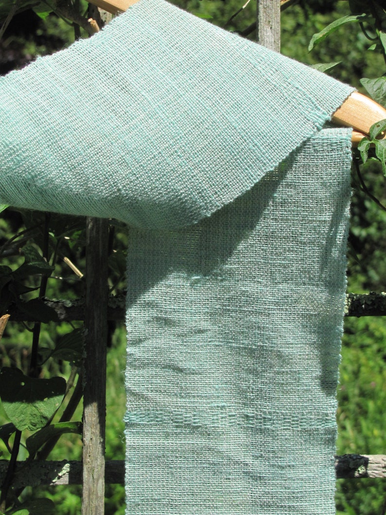 May include: A light blue woven fabric scarf hanging on a wooden hanger. The fabric has a textured, woven pattern.
