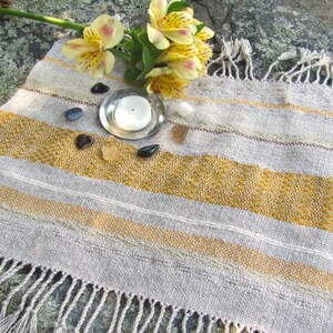 Small Altar Cloth Table Mat Centerpiece, Artisan Handmade Hand Woven Sun Yellow, Brown, Ivory White and Beige Gray Handcrafted by Amy C. Lund with striped bands of color and textural patterns and hand knotted fringe.  Styled with a small, white candle on a metal plate circled by small, colorful stones, and yellow freesia on a stone surface.