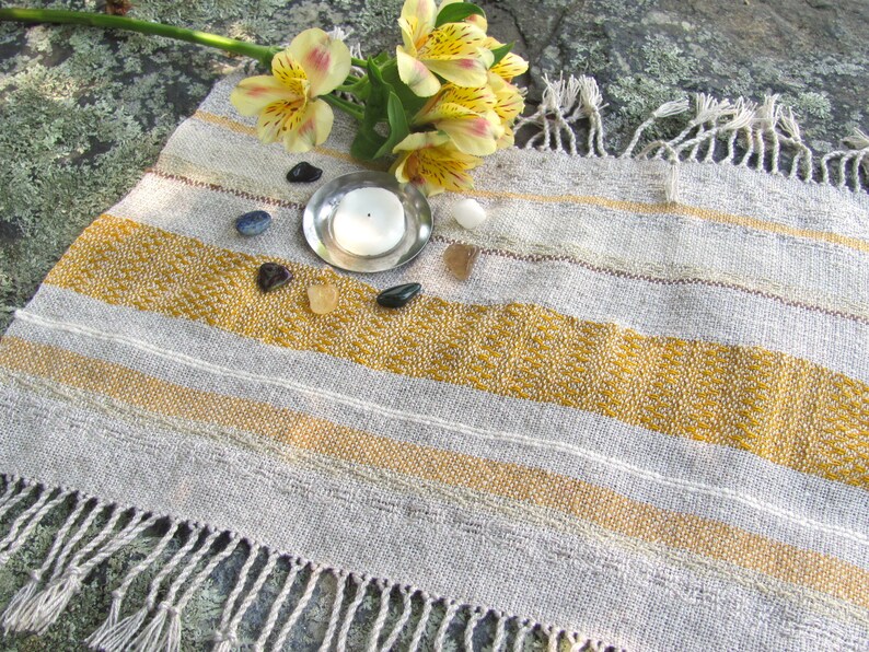 Small Altar Cloth Table Mat Centerpiece, Artisan Handmade Hand Woven Sun Yellow, Brown, Ivory White and Beige Gray Handcrafted by Amy C. Lund with striped bands of color and textural patterns and hand knotted fringe.  Styled with a small, white candle on a metal plate circled by small, colorful stones, and yellow freesia on a stone surface.