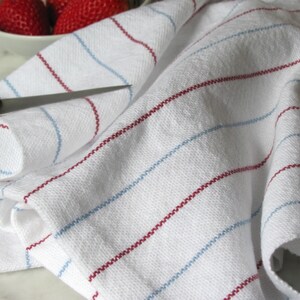 A white hand loom woven kitchen towel with red and blue stripes, shown as an example for Kitchen Tea Towels Weaving Pattern Draft PDF, Easy Beginner Handweaving 4H Harness Shaft Table or Floor Loom Digital Download Instructions as an artful Kitchenware Cookware Accessory for Gourmet Chef Home Baking, Cooking Designed by Master Weaver Amy C. Lund. The towel is folded and lying on a marble surface.