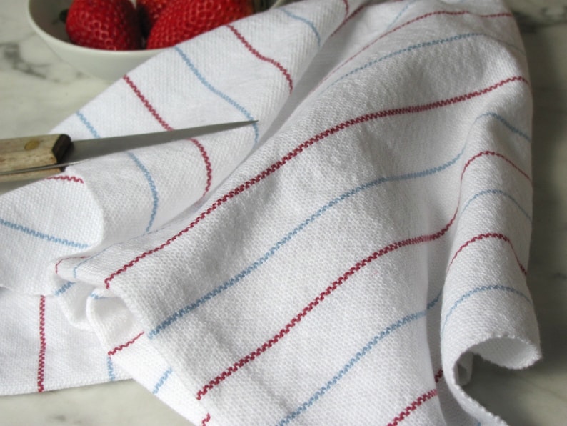 A white hand loom woven kitchen towel with red and blue stripes, shown as an example for Kitchen Tea Towels Weaving Pattern Draft PDF, Easy Beginner Handweaving 4H Harness Shaft Table or Floor Loom Digital Download Instructions as an artful Kitchenware Cookware Accessory for Gourmet Chef Home Baking, Cooking Designed by Master Weaver Amy C. Lund. The towel is folded and lying on a marble surface.