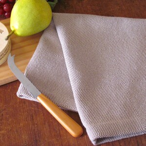 A brown and white Twill Block patterned hand woven towel, shown as an example for Table or Floor Loom Weaving Pattern Draft PDF, 4 H Harness Shaft Handweaving Cotton Kitchen Hand Woven Dish Tea Towel Downloadable Tutorial, an artful Kitchenware, Cookware Accessory for Gourmet Chef, Home Baking, Cooking, Instructions Designed by Master Weaver Amy C. Lund. Pictured with with a cheese knife, pear, grapes and cheese on a wooden cutting board.