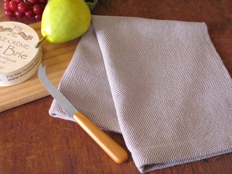 A brown and white Twill Block patterned hand woven towel, shown as an example for Table or Floor Loom Weaving Pattern Draft PDF, 4 H Harness Shaft Handweaving Cotton Kitchen Hand Woven Dish Tea Towel Downloadable Tutorial, an artful Kitchenware, Cookware Accessory for Gourmet Chef, Home Baking, Cooking, Instructions Designed by Master Weaver Amy C. Lund. Pictured with with a cheese knife, pear, grapes and cheese on a wooden cutting board.