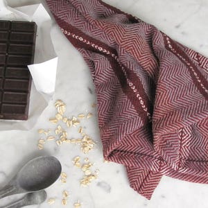 Handcrafted Kitchen Dish Towel, Gourmet Baking, Home Cooking gift for Chef, Baker, Cook, Foodie. An Eco Sustainable, Reusable, Artisan Handmade Cookware Kitchenware Accessory Hand Woven Cotton Towel by Amy C. Lund in Deep Burgundy Red, Beige, White & Fudge Brown Stripe Cloth for Rustic French Country or Modern Farmhouse Decor with alternating Chevron Diamond Zig Zag Herringbone. Styled on a white marble surface with a bar of chocolate, metal measuring spoons, and oats.