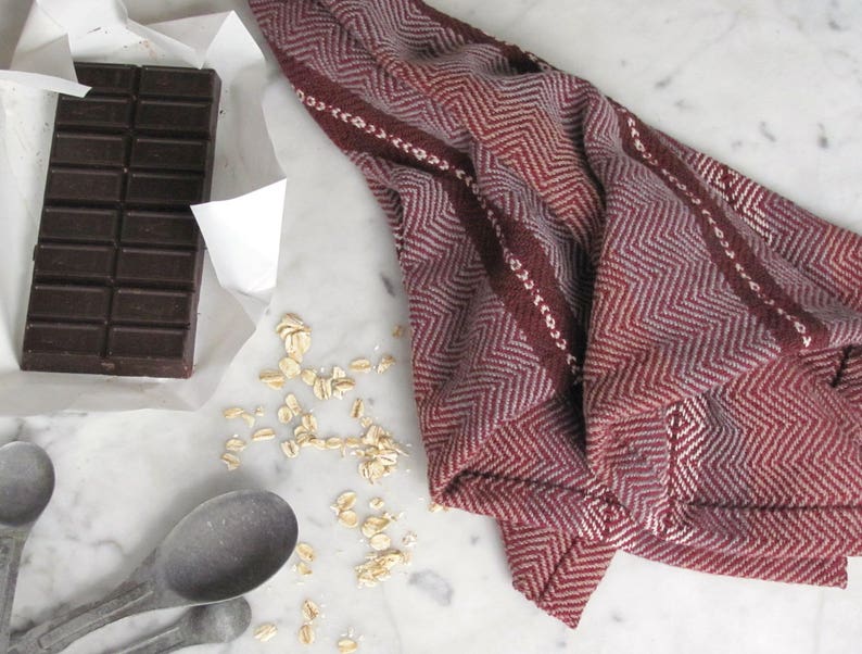 Handcrafted Kitchen Dish Towel, Gourmet Baking, Home Cooking gift for Chef, Baker, Cook, Foodie. An Eco Sustainable, Reusable, Artisan Handmade Cookware Kitchenware Accessory Hand Woven Cotton Towel by Amy C. Lund in Deep Burgundy Red, Beige, White & Fudge Brown Stripe Cloth for Rustic French Country or Modern Farmhouse Decor with alternating Chevron Diamond Zig Zag Herringbone. Styled on a white marble surface with a bar of chocolate, metal measuring spoons, and oats.