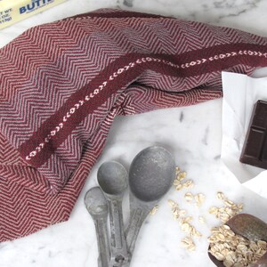 Handcrafted Kitchen Dish Towel, Gourmet Baking, Home Cooking gift for Chef, Baker, Cook, Foodie. An Eco Sustainable, Reusable, Artisan Handmade Cookware Kitchenware Accessory Hand Woven Cotton Towel by Amy C. Lund in Deep Burgundy Red, Beige, White & Fudge Brown Stripe Cloth for Rustic French Country or Modern Farmhouse Decor with alternating Chevron Diamond Zig Zag Herringbone. Styled on a white marble surface with a bar of chocolate, stick of butter, metal measuring spoons, and oats.