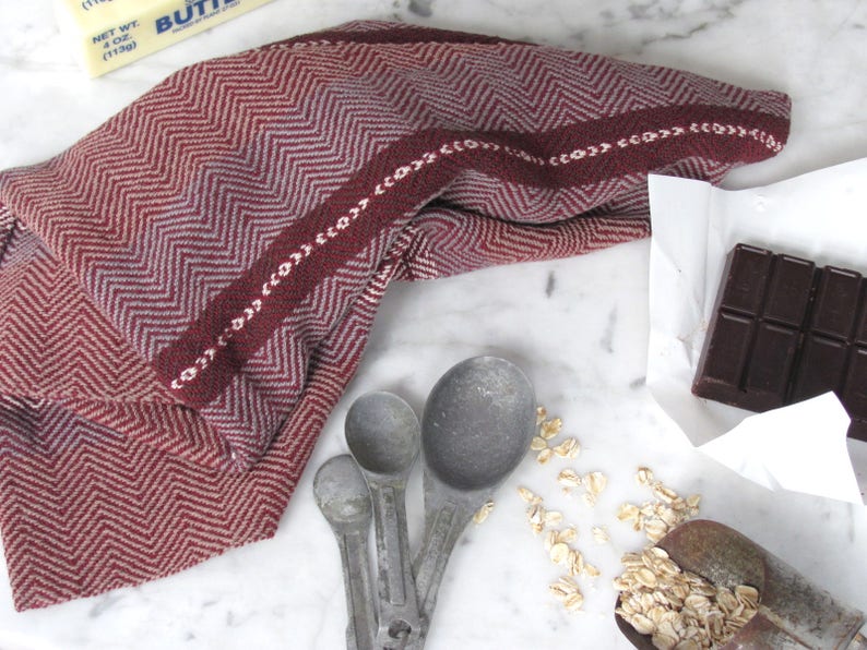 Handcrafted Kitchen Dish Towel, Gourmet Baking, Home Cooking gift for Chef, Baker, Cook, Foodie. An Eco Sustainable, Reusable, Artisan Handmade Cookware Kitchenware Accessory Hand Woven Cotton Towel by Amy C. Lund in Deep Burgundy Red, Beige, White & Fudge Brown Stripe Cloth for Rustic French Country or Modern Farmhouse Decor with alternating Chevron Diamond Zig Zag Herringbone. Styled on a white marble surface with a bar of chocolate, stick of butter, metal measuring spoons, and oats.