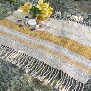 Small Altar Cloth Table Mat Centerpiece, Artisan Handmade Hand Woven Sun Yellow, Brown, Ivory White and Beige Gray Handcrafted by Amy C. Lund with striped bands of color and textural patterns and hand knotted fringe.  Styled with a small, white candle on a metal plate circled by small, colorful stones, and yellow freesia on a stone surface.