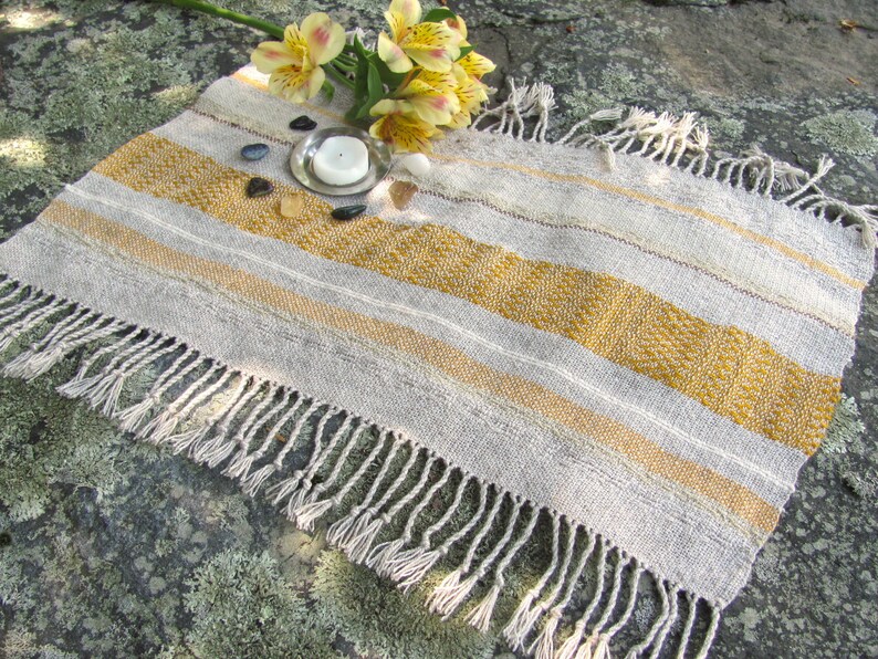 Small Altar Cloth Table Mat Centerpiece, Artisan Handmade Hand Woven Sun Yellow, Brown, Ivory White and Beige Gray Handcrafted by Amy C. Lund with striped bands of color and textural patterns and hand knotted fringe.  Styled with a small, white candle on a metal plate circled by small, colorful stones, and yellow freesia on a stone surface.