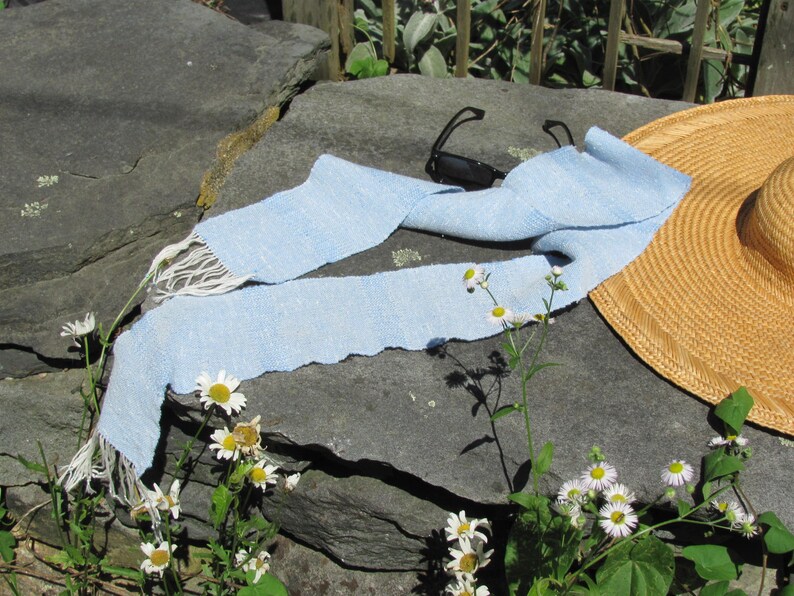 Artisan Handmade Light Blue Spring Summer Scarf, Minimalist Zen Serenity, Simplicity, Soothing Tranquil Tones Meditation Table Runner, Gender Neutral Unisex Men's or Women's Om Yoga, Spiritual Prayer Clothing, Hand Woven Cotton Scarf, Slow Fashion Accessory or Home Decor Accent Gift Idea. Styled casually on a gray rock with black sunglasses and white daisies, a straw hat is in the background.
