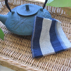 May include: A blue cast iron teapot with a bamboo design sits on a woven wicker tray with a blue and white striped tea towel.