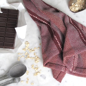 Handcrafted Kitchen Dish Towel, Gourmet Baking, Home Cooking gift for Chef, Baker, Cook, Foodie. An Eco Sustainable, Reusable, Artisan Handmade Cookware Kitchenware Accessory Hand Woven Cotton Towel by Amy C. Lund in Deep Burgundy Red, Beige, White & Fudge Brown Stripe Cloth for Rustic French Country or Modern Farmhouse Decor with alternating Chevron Diamond Zig Zag Herringbone. Styled on a white marble surface with a bar of chocolate, metal measuring spoons, and oats.
