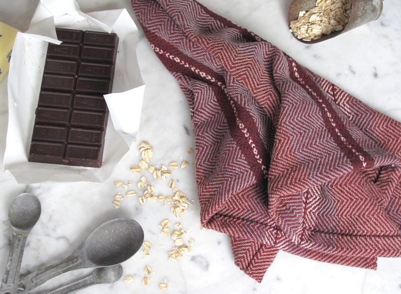 Handcrafted Kitchen Dish Towel, Gourmet Baking, Home Cooking gift for Chef, Baker, Cook, Foodie. An Eco Sustainable, Reusable, Artisan Handmade Cookware Kitchenware Accessory Hand Woven Cotton Towel by Amy C. Lund in Deep Burgundy Red, Beige, White & Fudge Brown Stripe Cloth for Rustic French Country or Modern Farmhouse Decor with alternating Chevron Diamond Zig Zag Herringbone. Styled on a white marble surface with a bar of chocolate, metal measuring spoons, and oats.