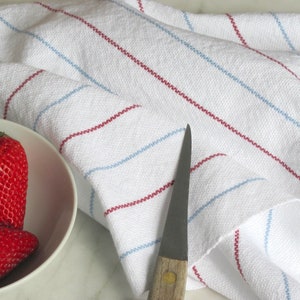 A hand woven white kitchen towel with red and blue stripes, shown as an example for Kitchen Tea Towels Weaving Pattern Draft PDF, Easy Beginner Handweaving 4H Harness Shaft Table or Floor Loom Digital Download Instructions as an artful Kitchenware Cookware Accessory for Gourmet Chef Home Baking, Cooking Designed by Master Weaver Amy C. Lund. There are three red strawberries in a white bowl next to the towel. A silver knife is also lying on the surface.