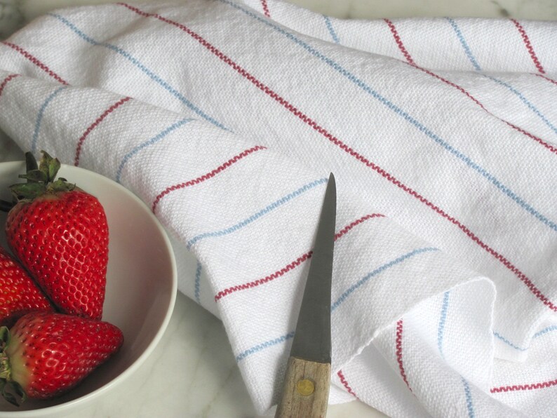 A hand woven white kitchen towel with red and blue stripes, shown as an example for Kitchen Tea Towels Weaving Pattern Draft PDF, Easy Beginner Handweaving 4H Harness Shaft Table or Floor Loom Digital Download Instructions as an artful Kitchenware Cookware Accessory for Gourmet Chef Home Baking, Cooking Designed by Master Weaver Amy C. Lund. There are three red strawberries in a white bowl next to the towel. A silver knife is also lying on the surface.