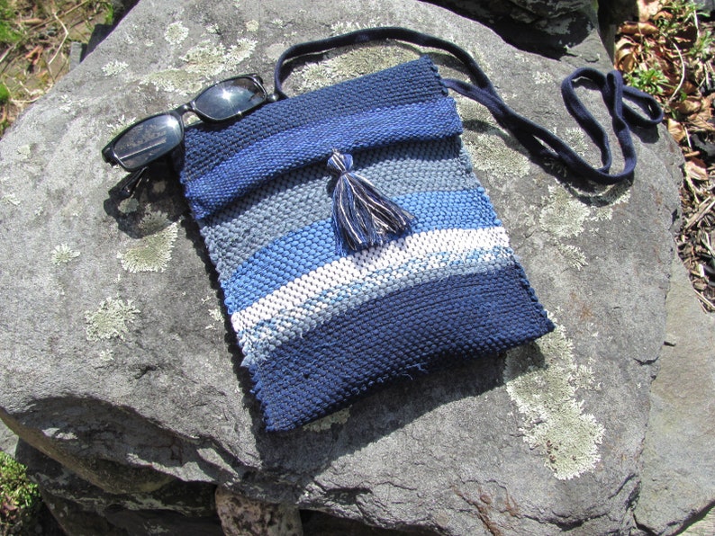 Small Crossbody Bag Wallet or Phone Purse is Handmade, Artisan Hand Woven in Indigo Navy Blue with Zero Waste, Eco Friendly Upcycled Cloth, Vegan Recycled Fabric. This Sustainable Slow Fashion Accessory Handbag, Boho Cash Change Pouch or Money Shoulder Pouch is Handcrafted by Amy C. Lund. Styled casually with black sunglasses on a gray rock.