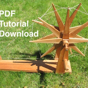 May include: A wooden spinning wheel with a starburst design and a white string wrapped around it. The wheel is sitting on a wooden base and is on a grassy surface. The text "PDF Tutorial Download" is in the upper left corner of the image.