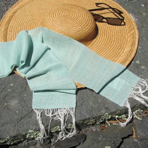 May include: A light blue woven scarf with white fringe, lying on a gray stone surface next to a straw hat and sunglasses.