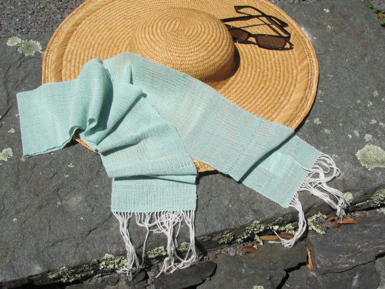 May include: A light blue woven scarf with white fringe, lying on a gray stone surface next to a straw hat and sunglasses.