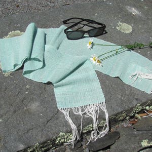 May include: A light blue woven scarf with white fringe, lying on a gray stone surface with white daisies and sunglasses.