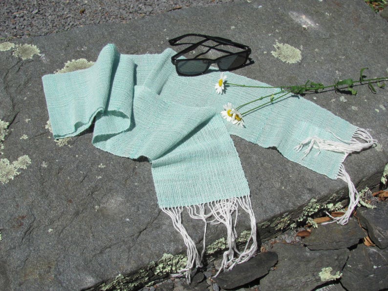 May include: A light blue woven scarf with white fringe, lying on a gray stone surface with white daisies and sunglasses.