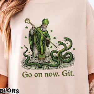 Puede incluir: Camiseta color marfil con un gráfico de un santo con túnica verde, sosteniendo un báculo y luchando contra serpientes verdes. El texto "Go on now. Git." está debajo. La camiseta está etiquetada como "Comfort Colors".