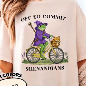 May include: Ivory-colored t-shirt with the text "OFF TO COMMIT SHENANIGANS." The graphic features a frog wizard riding a yellow bicycle with a cat in the basket. The shirt is from the Comfort Colors collection.