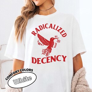 Op de afbeelding: Wit T-shirt met de rode tekst "RADICALIZED BY BASIC DECENCY" en een rode kolibrie-afbeelding. Het shirt heeft ook de woorden "COMFORT COLORS" en "White" in een witte ovaal.