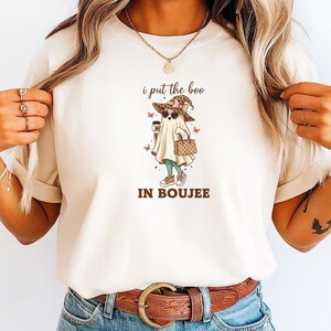 May include: Beige t-shirt with a graphic of a ghost wearing sunglasses, a hat, and carrying a coffee cup and purse. The text "i put the boo IN BOUJEE" is above and below the ghost illustration.