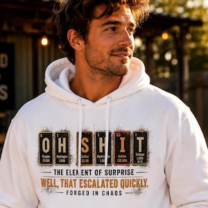 Könnte beinhalten: Ein weißer Kapuzenpullover mit einer Grafik aus chemischen Elementen, die OH SHIT buchstabieren. Unter den Elementen steht der Text THE ELEMENT OF SURPRISE, WELL, THAT ESCALATED QUICKLY und FORGED IN CHAOS in einer abgenutzten Schriftart.