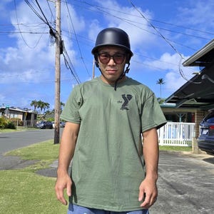 May include: An olive green t-shirt with a black graphic and the word "the" is worn with a black helmet and sunglasses. The person is standing outside on a sunny day.