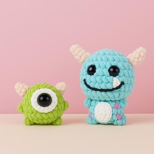 May include: Two handmade crochet monster toys. One is lime green with a large black eye and white accents. The other is light blue with two black eyes, a smile, and a white belly with pink accents. Both have small beige horns.