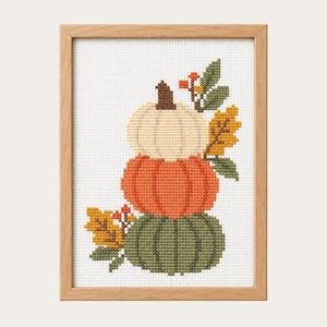 Puede incluir: Un bordado de punto de cruz en un marco de madera clara que muestra una pila vertical de tres calabazas en color crema, naranja y verde oscuro. El diseño está decorado con pequeñas bayas naranjas y hojas de otoño amarillas y verdes.