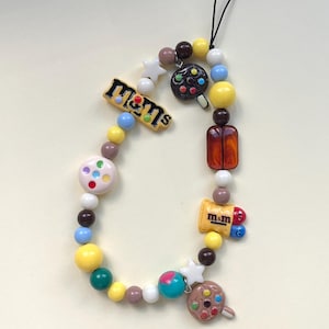 Op de afbeelding: Een kleurrijke telefoonbedel met diverse kralen en bedels. Het ontwerp bevat gele, blauwe, bruine en witte kralen, samen met bedels in de vorm van sterren, snoepjes en M&M's. Een zwart koord is bevestigd voor gemakkelijk dragen.