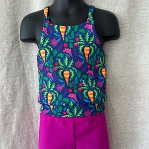 Puede incluir: Un conjunto de dos piezas para niña con una blusa sin mangas y pantalones cortos. La blusa tiene un fondo azul oscuro con un estampado colorido de zanahorias, guisantes y otras verduras. Los pantalones cortos son de color magenta sólido.