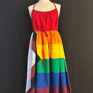 Op de afbeelding: Een rode halterjurk met een regenboogkleurige rok. De rok is gemaakt van verschillende gekleurde stofpanelen, elk met een ander patroon. De jurk is in de taille gebonden met een rood lint.